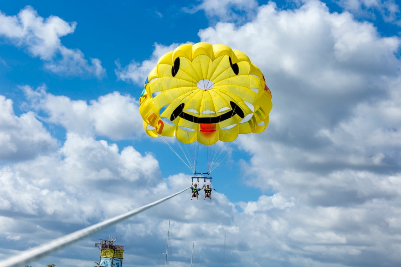Two people soaring under a yellow smiley-face parasail parachute against a blue sky in Cancun