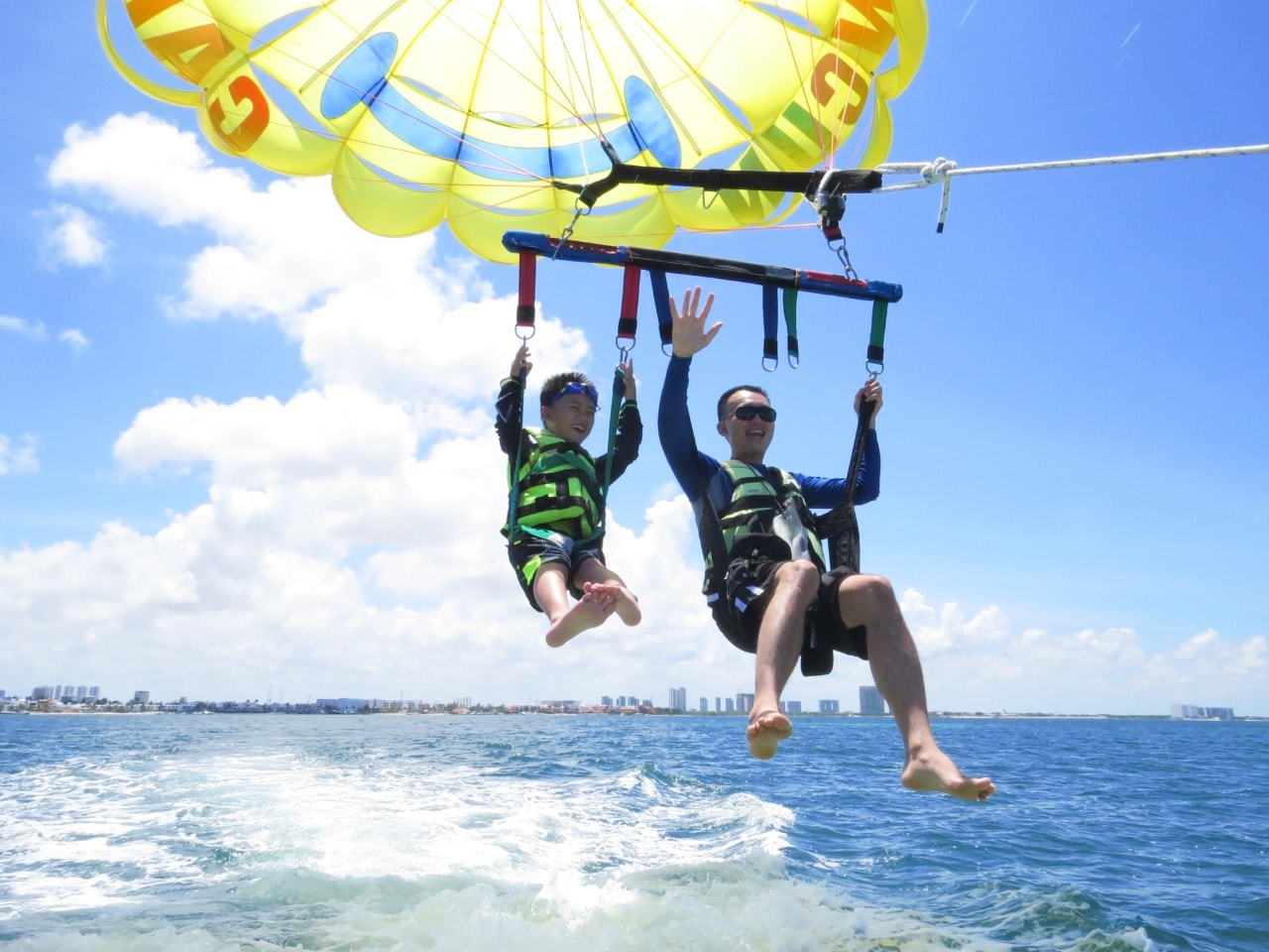 Father and son waving at the camera while parasailing just above the ocean waves in Cancun